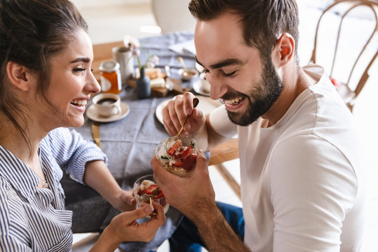 Image of attractive brunette couple eating panna cotta dessert together while sitting at table at home
