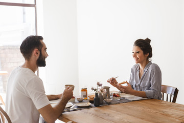 Image of caucasian brunette couple eating together at table while having breakfast in apartment