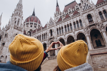 Obraz premium girl and friend in yellow caps holding hands in the shape of a heart against the background of the parliament in Budapest. I love Budapest. 