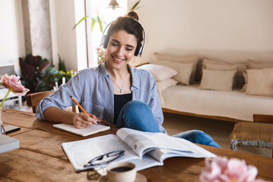 Image of smart charming woman listening to music while studying with exercise books at home