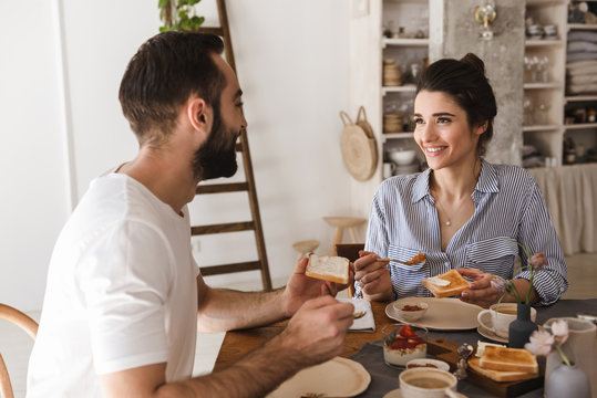 Image of lovely brunette couple eating together at table while having breakfast in apartment
