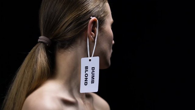 Male hand putting dumb blonde label on female ear, humiliation, stereotypes
