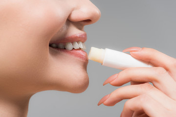 cropped view of cheerful girl holding lip balm near lips isolated on grey
