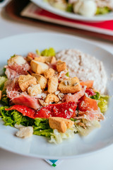 Plate of salad with tomatoes, jamon and baked vegetables on the white table. Glass of iced water with mint and lemon.