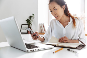 Smiling asian business woman using smartphone while sitting by table
