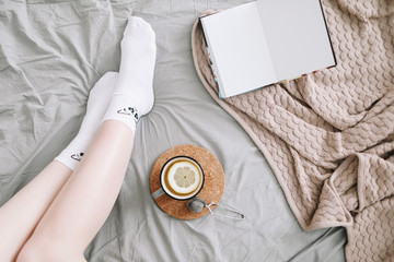 Cozy flatlay of woman legs in bed with a book and a cup of lemon tea. Cozy morning at home. 