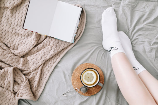 Cozy flatlay of woman legs in white stockings in bed with knitted sweater and book and a cup of lemon tea 