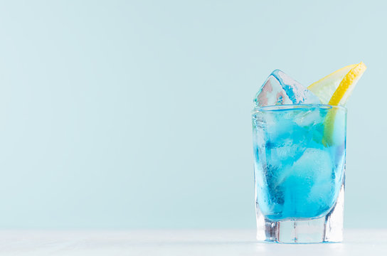 Alcohol juicy fruit blue cocktail with curacao liquor, lemon slice, ice cubes in frozen shot glass in modern elegant pastel blue interior.