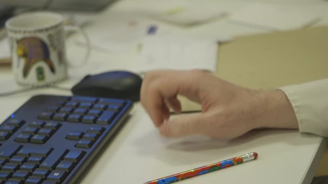 Handheld, medium close up of a hand tapping impatiently on a desk. A colorful pencil and a computer also sit on the desk.