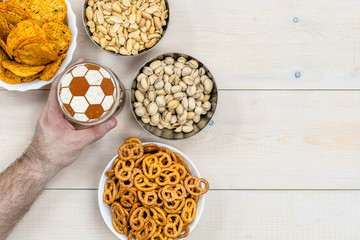 Man's hand holding a mug of beer with a soccer ball on a beer foam and  snacks on a light wooden background. Top view. Empty space for text