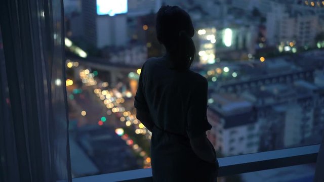 Silhouette of woman looking at city at night, standing by window at home