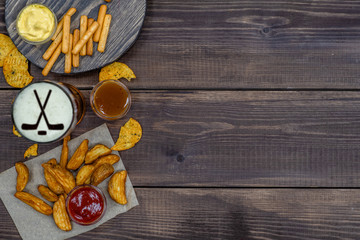 Diverse snacks and beer with silhouettes of hockey sticks on beer foam on dark wooden background. Top view. Empty space for text