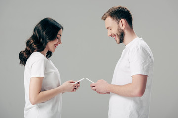 side view of cheerful man and woman in white t-shirts using smartphones isolated on grey