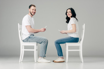 smiling man and woman in white t-shirts and blue jeans sitting on chairs and using smartphones on grey background