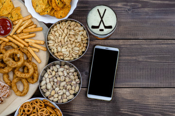 Mix of snacks and beer with silhouettes of hockey sticks on beer foam, and smartpnone on dark wooden background. Internet delivery concept. Top view. Empty space for text