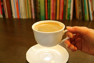 Female's Hand Holding White Cup of Hot Coffee with Blurry Rows of Books in Background 