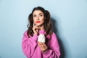 Portrait of doubtful young woman with two ponytails smiling while eating sweet cupcake