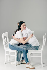 Obraz premium young couple embracing while sitting on white chairs and using smartphones on grey background