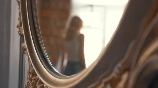 Close-up of mirror reflection of young blond woman in white t-shirt and blue jeans walking near a window in her appartments. Action. Relaxing at home