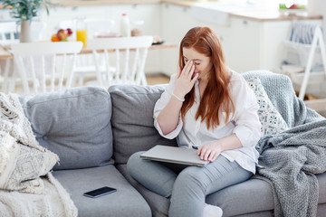 Tired red-haired woman closing laptop and rubbing nose bridge trying to concentrate and relieve fatigue sitting on sofa at home