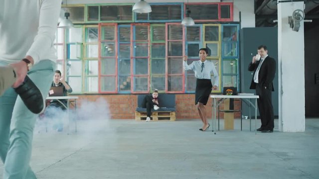 Two colleagues ride in an office chair. The manager lay down on the chair like a superman, and his partner rolls him merrily around the office. Smoke screen. Office life. Business team. Celebration