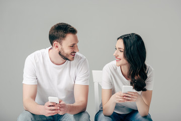 smiling couple looking at each other while sitting on chairs and using smartphone isolated on grey