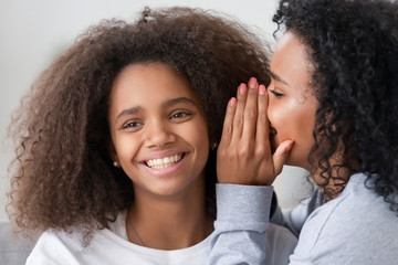 African American mother whispering to smiling teen daughter ear - obrazy, fototapety, plakaty
