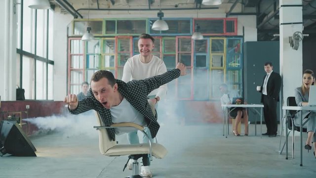 Young businessman is riding a chair in the office like a superman. His colleague helps him. Smoke screen. Office life. Business team. Celebration