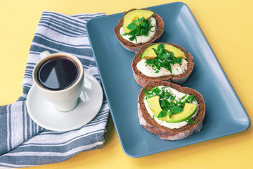 Cup of coffee and plate with avocado sandwiches on a yellow table, healthy breakfast, top view