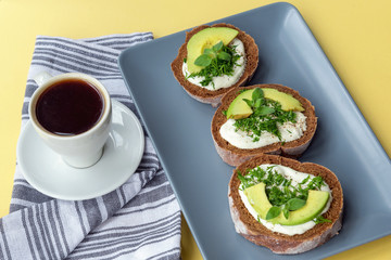 Cup of coffee and plate with avocado sandwiches on a yellow table, healthy breakfast, top view