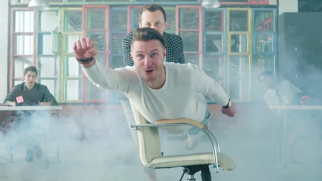The manager is riding a chair in the office like a superman. His colleague helps him. Office entertainment. Smoke screen. Office life. Business team. Slow mo