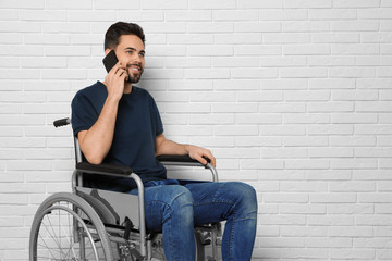 Fototapeta na wymiar Young man in wheelchair talking on phone near brick wall indoors. Space for text
