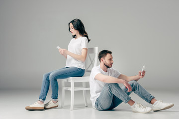 concentrated woman using smartphone near focused man sitting on floor with smartphone on grey background