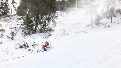 Fototapeta na wymiar Snowgun spray artificial ice crystals over ski piste, snowmaking in winter sports resort, trees in background