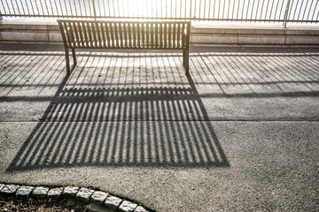 Morning sun cast lines of shadows on asphalt pavement through fence and empty bench by the river
