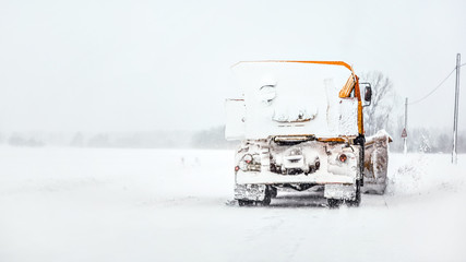 Orange plough truck on snow covered road, gray sky and trees in background, view from car driving...