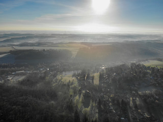 Aerial view of foggy and cold winter morning with blue sky facing the sun over the forest and farmland in Belgium, Walloon Brabant