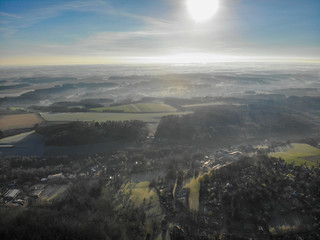 Aerial view of foggy and cold winter morning with blue sky facing the sun over the forest and farmland in Belgium, Walloon Brabant © Unwind