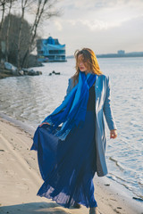 Gorgeous european woman in warm coat and dress on a walk in park near river. Windy weather. Her clothes fly in the wind. Sad, sensual and thughtful expression on her face