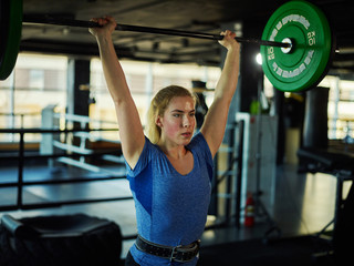 Waist-up portrait of strong young woman lifting barbell with weight plates overhead during workout...