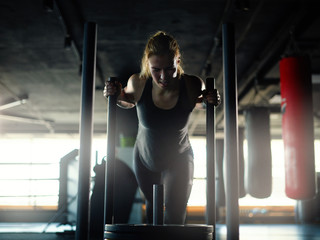 Fototapeta na wymiar Backlit shot of determined young woman making great effort to pull weight sled with plates during weight loss workout in gym..