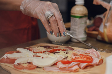 A woman in disposable gloves sprinkles cheese pizza with ham and tomatoes. Cooking pizza