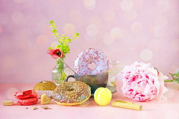 A black cup of coffee, donuts in colored glaze, a bunch of flowers, macaroons, cinnamon sticks, sugar bowl, cakes stand on a pink background. The concept of a sweet breakfast, save space.