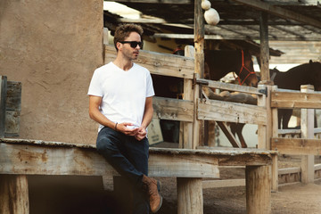 Young handsome bearded man is staying on a western city background surrounded by wooden fence. Guy is wearing a white empty t-shirt without logo. Horizontal mock-up style.