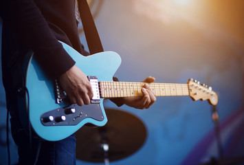 Naklejka na ściany i meble A man in a black sweater in blue jeans plays a blue electric guitar, illuminated by sunlight, on a blue background.