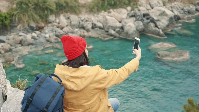 Tourist girl sitting on the edge of a cliff in the lagoon and shooting selfie with a beautiful view on background. Cloudy autumn day. 4K