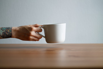 hand with tattoos holding a white mug on the table on a light background