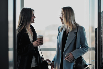 Two beautiful girls, a blonde and a brunette, are wearing a coat in a glass elevator with a window....