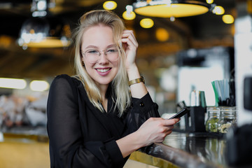 A young beautiful blond woman with glasses sits at the bar in a yellow interior. Drinks alcoholic cocktail with a straw and green mint.