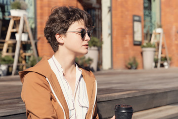 Young beautiful girl with haircut sitting on the street against a red brick wall and drinks coffee.